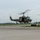Three olive drab military helicopters, identifiable as UH-1 'Huey' type aircraft, are captured in low-level flight over an airport tarmac. The helicopters appear to be performing a formation flight or demonstration, likely at an airshow or military event. Military unit insignia and markings are visible on the fuselages.