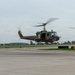 Three olive drab military helicopters, identifiable as UH-1 'Huey' type aircraft, are captured in low-level flight over an airport tarmac. The helicopters appear to be performing a formation flight or demonstration, likely at an airshow or military event. Military unit insignia and markings are visible on the fuselages.