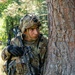 A U.S. Army soldier in full combat gear is taking cover behind a large tree trunk in a wooded training environment. The soldier is wearing OCP (Operational Camouflage Pattern) uniform and helmet with identifier '2CG9077' visible, and is holding an M4-style carbine rifle. The image appears to depict a field training exercise based on the MILES/training equipment visible on the weapon and helmet.