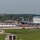 An aerial view of a military air show at what appears to be a U.S. Air Force base, with large crowds gathered along the flight line. At least two large military aircraft, including what appears to be a C-5 Galaxy or similar heavy transport aircraft marked 'MT', are on static display. White vendor tents and spectator areas are set up along the tarmac with hundreds of attendees visible.