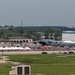 An aerial view of a military air show at what appears to be a U.S. Air Force base, with large crowds gathered along the flight line. At least two large military aircraft, including what appears to be a C-5 Galaxy or similar heavy transport aircraft marked 'MT', are on static display. White vendor tents and spectator areas are set up along the tarmac with hundreds of attendees visible.