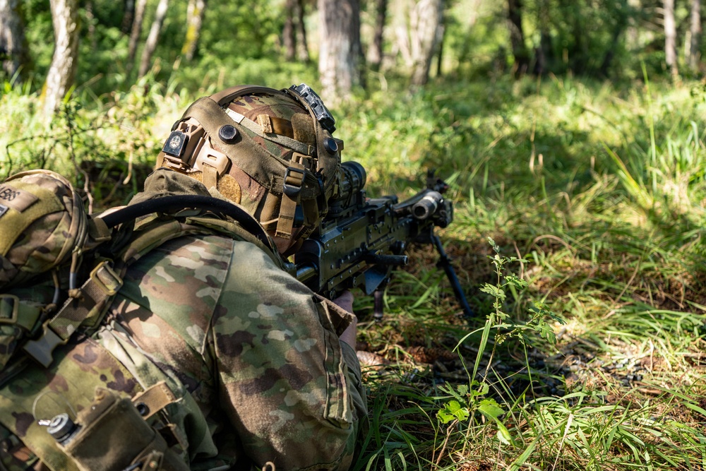 A soldier in full combat gear is in a prone firing position in a forested environment, aiming a light machine gun equipped with a bipod. The individual is wearing multicam camouflage uniform and a tactical helmet with mounted accessories. The setting appears to be a training exercise or operational patrol in a woodland area.