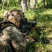 A soldier in full combat gear is in a prone firing position in a forested environment, aiming a light machine gun equipped with a bipod. The individual is wearing multicam camouflage uniform and a tactical helmet with mounted accessories. The setting appears to be a training exercise or operational patrol in a woodland area.