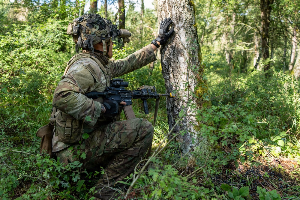 A soldier in full combat gear is crouching behind a tree in a forested environment, appearing to take cover or conduct reconnaissance. The individual is equipped with modern military equipment including a helmet-mounted night vision device, tactical vest, and an assault rifle. The setting suggests a field training exercise or operational patrol in a woodland environment.
