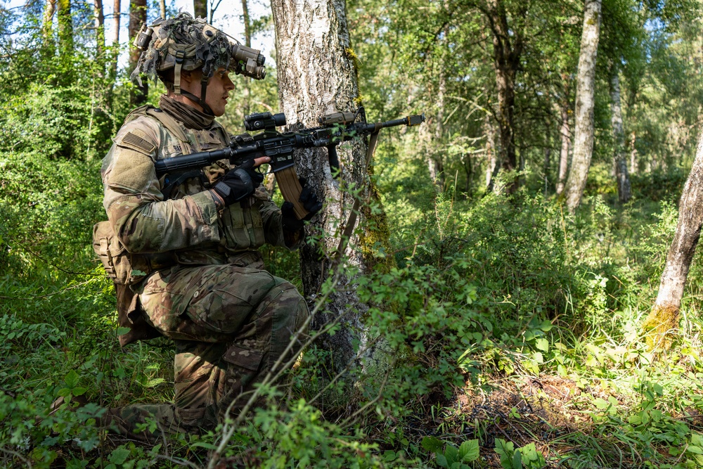 A soldier in full combat gear is positioned behind a tree in a forested environment, aiming a rifle in a tactical kneeling stance. The soldier is equipped with modern military equipment including a helmet-mounted night vision device, body armor, and an optical scope on the weapon. The setting appears to be a training exercise or patrol in a temperate woodland environment.