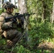 A soldier in full combat gear is positioned behind a tree in a forested environment, aiming a rifle in a tactical kneeling stance. The soldier is equipped with modern military equipment including a helmet-mounted night vision device, body armor, and an optical scope on the weapon. The setting appears to be a training exercise or patrol in a temperate woodland environment.