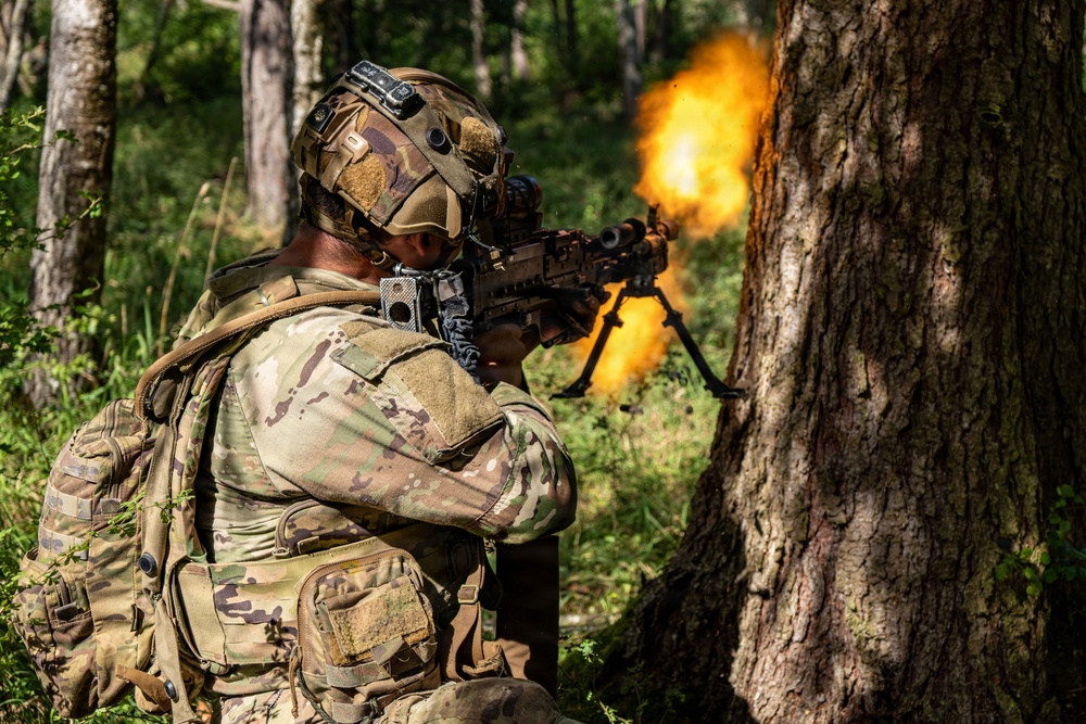 A soldier in full multicam camouflage gear is firing a light machine gun equipped with a bipod from a position beside a large tree in a forested environment. A large muzzle flash/fireball is visible at the barrel of the weapon, indicating active discharge. The image appears to document a military training exercise or combat operation.