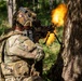 A soldier in full multicam camouflage gear is firing a light machine gun equipped with a bipod from a position beside a large tree in a forested environment. A large muzzle flash/fireball is visible at the barrel of the weapon, indicating active discharge. The image appears to document a military training exercise or combat operation.