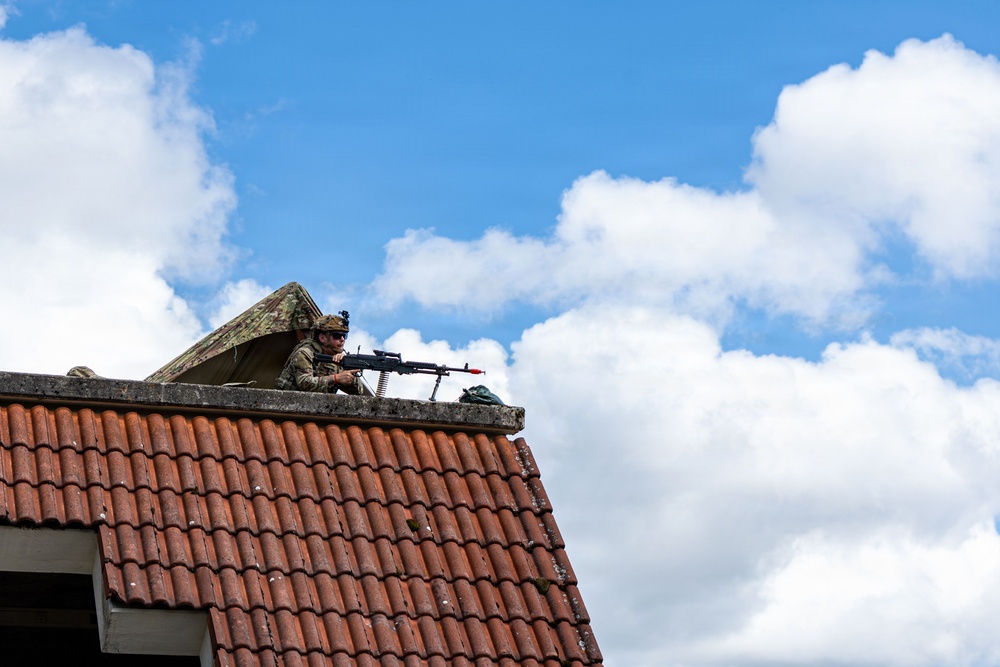 A soldier in camouflage uniform and helmet is positioned on a rooftop, aiming a machine gun with an ammunition belt visible. The soldier appears to be in a tactical overwatch position on a building with red clay roof tiles. The setting suggests a military exercise or operational deployment in a European-style built environment.