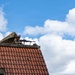A soldier in camouflage uniform and helmet is positioned on a rooftop, aiming a machine gun with an ammunition belt visible. The soldier appears to be in a tactical overwatch position on a building with red clay roof tiles. The setting suggests a military exercise or operational deployment in a European-style built environment.