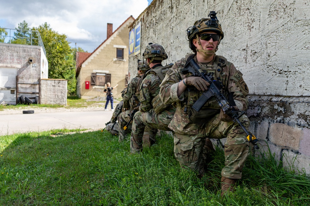 A group of soldiers in MultiCam/OCP camouflage uniforms are conducting what appears to be a military training exercise in a European village setting. The soldiers are positioned along a concrete wall in a tactical formation, armed with rifles and wearing full combat gear including helmets with night-vision mounts. A civilian figure is visible in the background, suggesting this may be a simulated urban operations or MOUT (Military Operations on Urban Terrain) exercise.