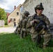 A group of soldiers in MultiCam/OCP camouflage uniforms are conducting what appears to be a military training exercise in a European village setting. The soldiers are positioned along a concrete wall in a tactical formation, armed with rifles and wearing full combat gear including helmets with night-vision mounts. A civilian figure is visible in the background, suggesting this may be a simulated urban operations or MOUT (Military Operations on Urban Terrain) exercise.
