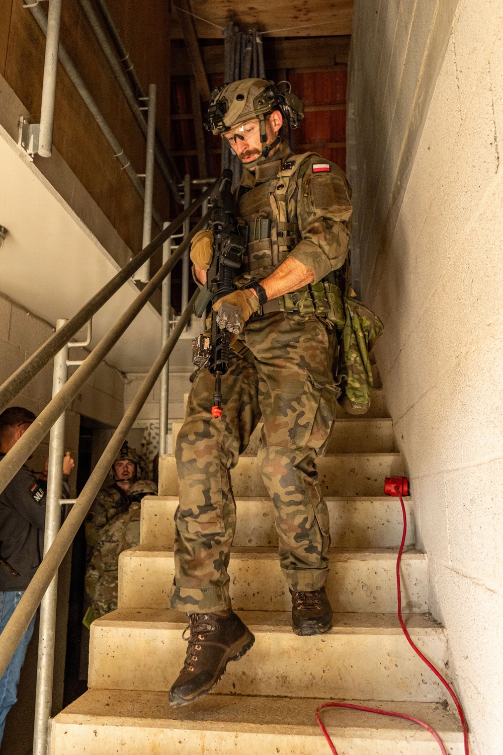A Polish soldier in full combat gear descends a concrete stairwell inside a building, holding an assault rifle. He wears a distinctive Polish woodland camouflage (wz. 93 pattern) uniform with a Polish flag patch visible on his left shoulder. Additional personnel in different camouflage uniforms are visible in the background at the base of the stairs.