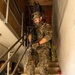 A Polish soldier in full combat gear descends a concrete stairwell inside a building, holding an assault rifle. He wears a distinctive Polish woodland camouflage (wz. 93 pattern) uniform with a Polish flag patch visible on his left shoulder. Additional personnel in different camouflage uniforms are visible in the background at the base of the stairs.