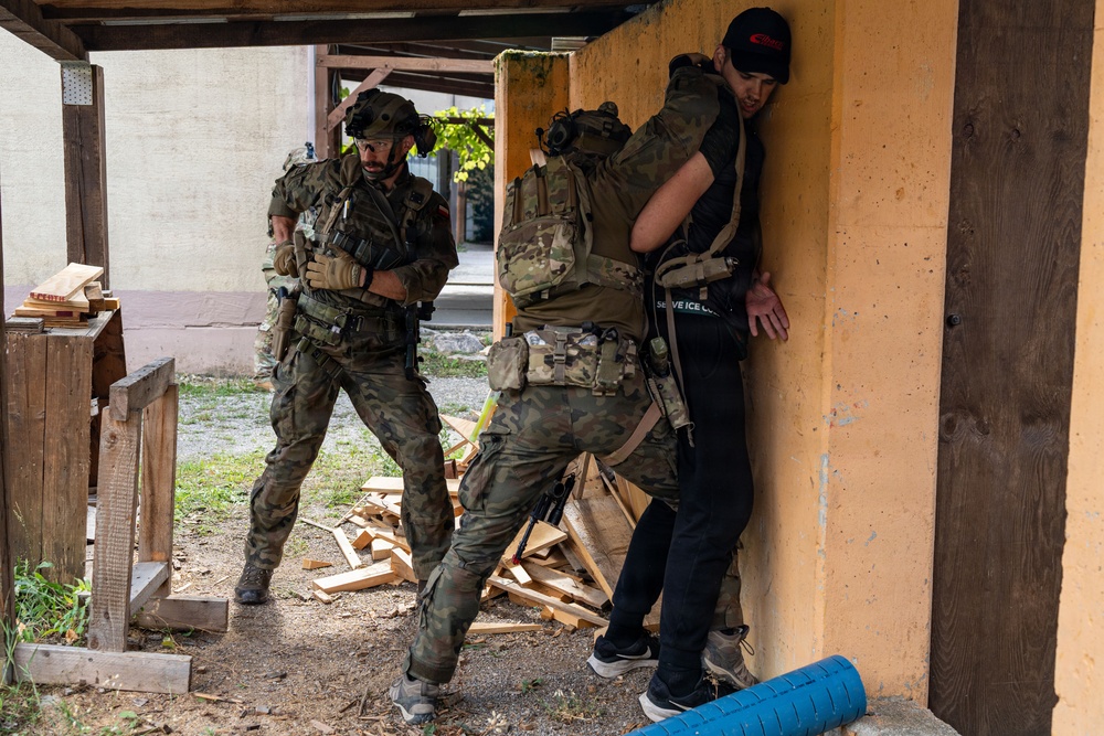 Two soldiers in camouflage uniforms and tactical gear are conducting what appears to be a military training exercise with a civilian role-player. The soldiers are performing a detainee handling or close-quarters combat drill near a building corner in a training environment. Scattered lumber and training props in the background suggest a purpose-built urban operations training facility.