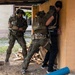 Two soldiers in camouflage uniforms and tactical gear are conducting what appears to be a military training exercise with a civilian role-player. The soldiers are performing a detainee handling or close-quarters combat drill near a building corner in a training environment. Scattered lumber and training props in the background suggest a purpose-built urban operations training facility.