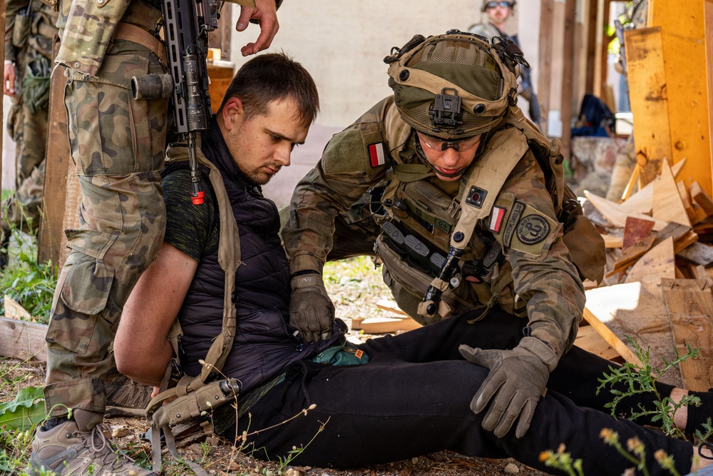 A soldier wearing Polish military insignia (red and white flag patches) is providing medical assistance or conducting a search of a seated civilian in a debris-filled environment. A second armed soldier stands nearby in camouflage uniform. The setting appears to be a military training exercise based on the controlled nature of the scene.