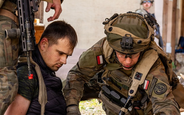 A soldier wearing Polish military insignia (red and white flag patches) is providing medical assistance or conducting a search of a seated civilian in a debris-filled environment. A second armed soldier stands nearby in camouflage uniform. The setting appears to be a military training exercise based on the controlled nature of the scene.