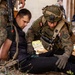 A soldier wearing Polish military insignia (red and white flag patches) is providing medical assistance or conducting a search of a seated civilian in a debris-filled environment. A second armed soldier stands nearby in camouflage uniform. The setting appears to be a military training exercise based on the controlled nature of the scene.