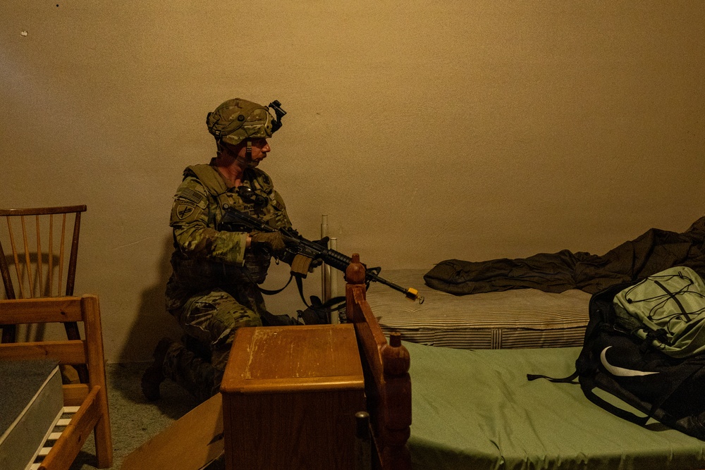 A soldier in full combat gear, including helmet with night vision mount and multicam uniform, is kneeling in a dimly lit room holding a rifle with a yellow training/blank firing attachment on the muzzle. The setting appears to be an interior room with a bed, wooden furniture, and military gear, suggesting a training exercise or clearing operation. The low ambient lighting and warm orange tone indicate either artificial indoor lighting or nighttime conditions.