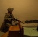 A soldier in full combat gear, including helmet with night vision mount and multicam uniform, is kneeling in a dimly lit room holding a rifle with a yellow training/blank firing attachment on the muzzle. The setting appears to be an interior room with a bed, wooden furniture, and military gear, suggesting a training exercise or clearing operation. The low ambient lighting and warm orange tone indicate either artificial indoor lighting or nighttime conditions.