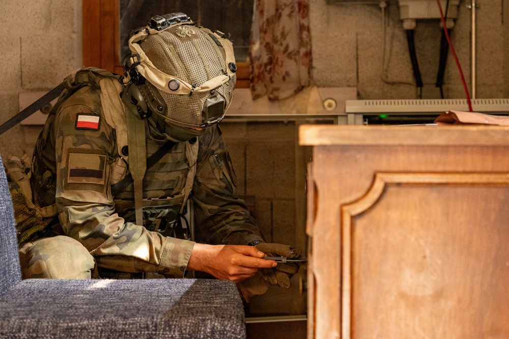 A fully equipped soldier wearing Polish military insignia and a tactical helmet is crouched in an indoor environment, examining a small handheld device. The soldier is dressed in camouflage fatigues with a Polish flag patch visible on the left shoulder. The setting appears to be a building interior, possibly during a training exercise or operational scenario.