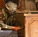 A fully equipped soldier wearing Polish military insignia and a tactical helmet is crouched in an indoor environment, examining a small handheld device. The soldier is dressed in camouflage fatigues with a Polish flag patch visible on the left shoulder. The setting appears to be a building interior, possibly during a training exercise or operational scenario.