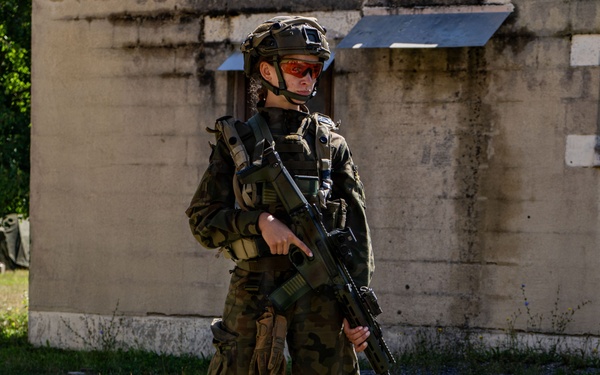 A fully equipped soldier in camouflage uniform stands guard over a seated civilian role-player during what appears to be a military training exercise. The soldier is armed with a rifle featuring a red training/blank-firing adapter on the muzzle, indicating a simulated scenario. The setting is an outdoor urban training environment with a concrete structure in the background.