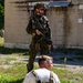A fully equipped soldier in camouflage uniform stands guard over a seated civilian role-player during what appears to be a military training exercise. The soldier is armed with a rifle featuring a red training/blank-firing adapter on the muzzle, indicating a simulated scenario. The setting is an outdoor urban training environment with a concrete structure in the background.