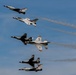 Four U.S. Air Force Thunderbirds demonstration team aircraft are flying in a diagonal echelon formation against a clear blue sky. The jets, identifiable as F-16 Fighting Falcons with their distinctive red, white, and blue livery, are producing white smoke trails as they perform. The image captures a precision aerial demonstration with the aircraft in tight, evenly-spaced formation.