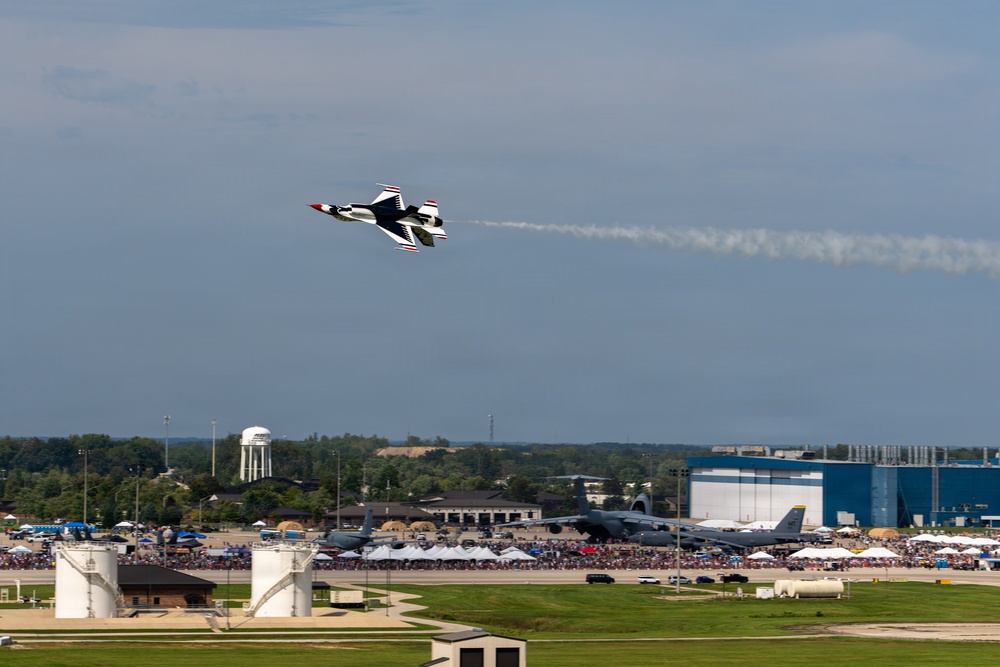 An air show is taking place at a military airbase, with a U.S. Air Force Thunderbirds F-16 jet performing an aerobatic maneuver while leaving a white smoke trail. Large crowds of spectators line the flight line below, with several large military aircraft visible on the ground. A prominent blue hangar building and a water tower are visible in the background.