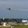 An air show is taking place at a military airbase, with a U.S. Air Force Thunderbirds F-16 jet performing an aerobatic maneuver while leaving a white smoke trail. Large crowds of spectators line the flight line below, with several large military aircraft visible on the ground. A prominent blue hangar building and a water tower are visible in the background.