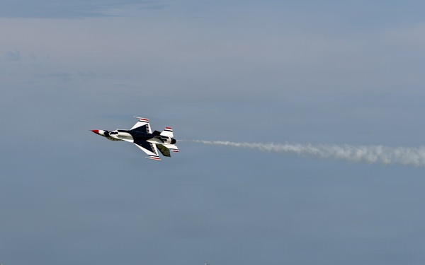An air show is taking place at a military airbase, with a U.S. Air Force Thunderbirds F-16 jet performing an aerobatic maneuver while leaving a white smoke trail. Large crowds of spectators line the flight line below, with several large military aircraft visible on the ground. A prominent blue hangar building and a water tower are visible in the background.