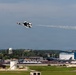 An air show is taking place at a military airbase, with a U.S. Air Force Thunderbirds F-16 jet performing an aerobatic maneuver while leaving a white smoke trail. Large crowds of spectators line the flight line below, with several large military aircraft visible on the ground. A prominent blue hangar building and a water tower are visible in the background.
