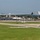 An airshow is taking place at a military or civilian airfield, featuring two jet aircraft performing a close formation or crossing maneuver at low altitude. A large crowd and extensive parking area are visible along the flight line in the background. The scene includes airfield infrastructure, buildings, a water tower, and a white bus on a taxiway.