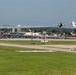 An airshow is taking place at a military or civilian airfield, featuring two jet aircraft performing a close formation or crossing maneuver at low altitude. A large crowd and extensive parking area are visible along the flight line in the background. The scene includes airfield infrastructure, buildings, a water tower, and a white bus on a taxiway.