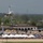 An air show is taking place at a military airbase, with a jet aircraft performing an inverted or banking maneuver at low altitude above the runway. A large crowd of spectators lines the flight line beneath white vendor tents, watching the performance. Additional large military transport aircraft are visible parked on the tarmac in the background.