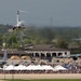 An air show is taking place at a military airbase, with a jet aircraft performing an inverted or banking maneuver at low altitude above the runway. A large crowd of spectators lines the flight line beneath white vendor tents, watching the performance. Additional large military transport aircraft are visible parked on the tarmac in the background.
