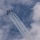 A formation of military jet aircraft fly in tight delta/diamond formation against a partly cloudy sky, leaving parallel white smoke or contrail streams behind them. The aircraft appear to be performing an aerobatic demonstration, consistent with a precision flight team display. The trails converge toward the lower center of the frame, creating a dramatic visual effect.