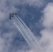 A formation of military jet aircraft fly in tight delta/diamond formation against a partly cloudy sky, leaving parallel white smoke or contrail streams behind them. The aircraft appear to be performing an aerobatic demonstration, consistent with a precision flight team display. The trails converge toward the lower center of the frame, creating a dramatic visual effect.