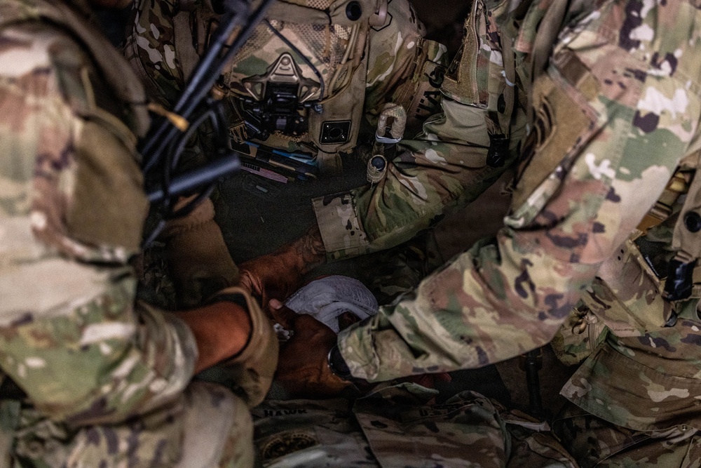 The image shows multiple military personnel in OCP (Operational Camouflage Pattern) uniforms gathered closely together, appearing to perform a tactical combat casualty care (TCCC) or first aid procedure. At least two soldiers are working with their hands on what appears to be a bandage or medical dressing being applied to a limb. The close-up framing and depth of field suggest this may be a training exercise or field medical response.