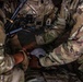 The image shows multiple military personnel in OCP (Operational Camouflage Pattern) uniforms gathered closely together, appearing to perform a tactical combat casualty care (TCCC) or first aid procedure. At least two soldiers are working with their hands on what appears to be a bandage or medical dressing being applied to a limb. The close-up framing and depth of field suggest this may be a training exercise or field medical response.