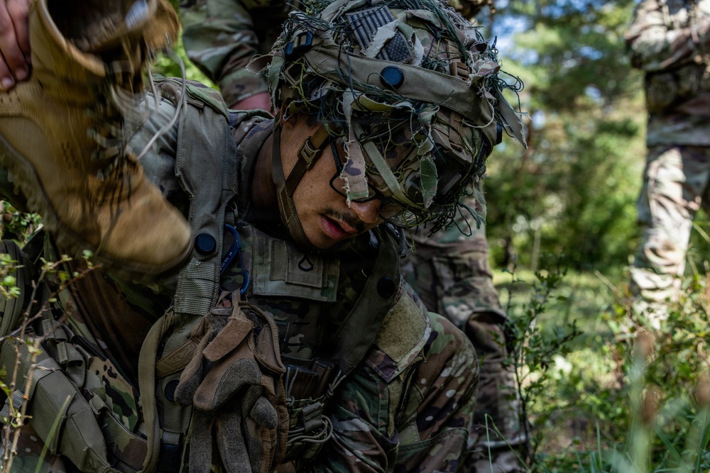 A soldier in full multicam combat gear and a camouflage-covered helmet is crouched down in a wooded outdoor environment. The soldier appears to be in a training or field exercise scenario, with another individual's gloved hand visible in the upper left and a second soldier partially visible on the right. The setting suggests a tactical field training exercise in a forested area.