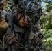 A soldier in full multicam combat gear and a camouflage-covered helmet is crouched down in a wooded outdoor environment. The soldier appears to be in a training or field exercise scenario, with another individual's gloved hand visible in the upper left and a second soldier partially visible on the right. The setting suggests a tactical field training exercise in a forested area.