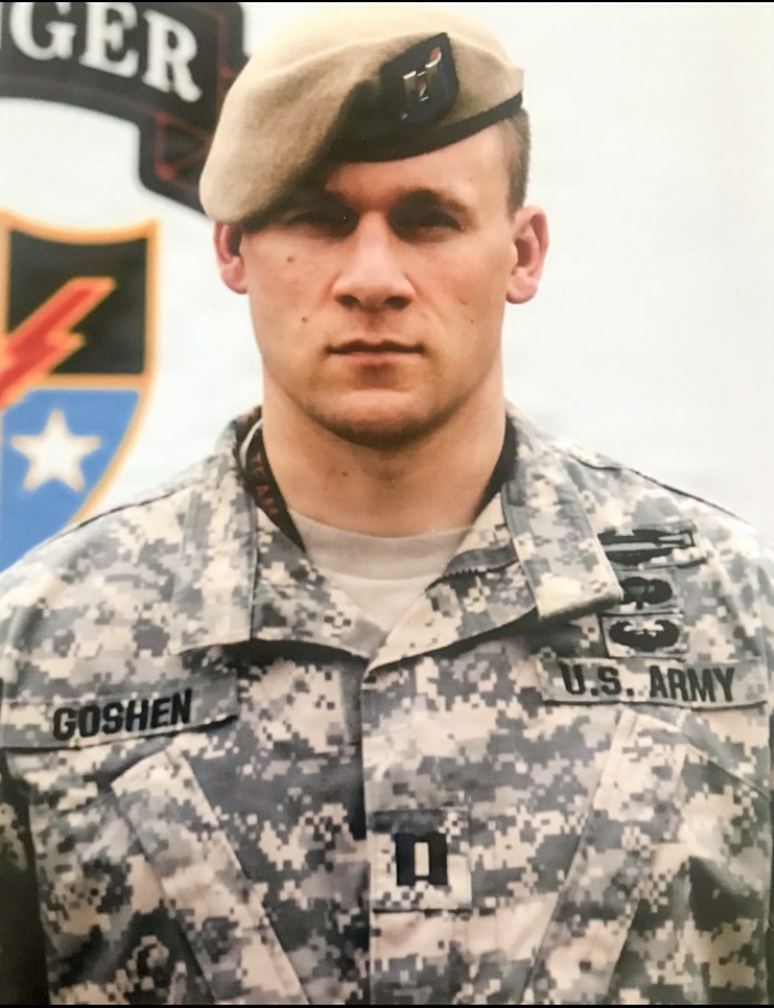 A U.S. Army soldier in Army Combat Uniform (ACU) digital camouflage pattern poses for an official military portrait. The soldier wears a tan beret with a flash insignia and has a name tape reading 'GOSHEN' on the left chest. The background displays what appears to be a Ranger training or unit insignia backdrop.