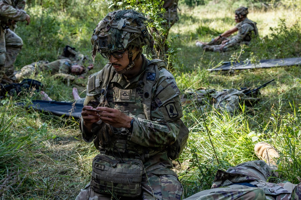 A U.S. Army soldier in full combat gear, including a camouflage-netted helmet with what appears to be a night-vision mount, is kneeling in a wooded field area while examining a small object or card in his hands. Several other soldiers are visible in the background, some lying on litters (stretchers) on the ground, suggesting a combat casualty care or MEDEVAC training exercise. The setting appears to be an outdoor field training environment.