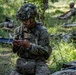 A U.S. Army soldier in full combat gear, including a camouflage-netted helmet with what appears to be a night-vision mount, is kneeling in a wooded field area while examining a small object or card in his hands. Several other soldiers are visible in the background, some lying on litters (stretchers) on the ground, suggesting a combat casualty care or MEDEVAC training exercise. The setting appears to be an outdoor field training environment.