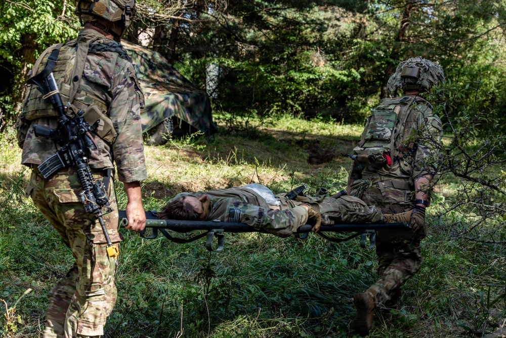 Two soldiers in camouflage uniforms are carrying a third soldier on a litter/stretcher through a wooded outdoor area. The casualty appears to be in full combat gear and is lying flat on the stretcher. This appears to be a military medical evacuation exercise or training scenario based on the controlled setting.