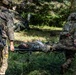 Two soldiers in camouflage uniforms are carrying a third soldier on a litter/stretcher through a wooded outdoor area. The casualty appears to be in full combat gear and is lying flat on the stretcher. This appears to be a military medical evacuation exercise or training scenario based on the controlled setting.
