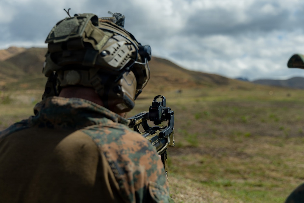 A military service member in full combat gear is seen from behind, aiming a rifle with an optical sight across an open, hilly terrain. The individual is wearing MARPAT (Marine pattern) camouflage uniform and a tactical helmet equipped with night vision device mounts. The setting appears to be an outdoor training range or field exercise environment.